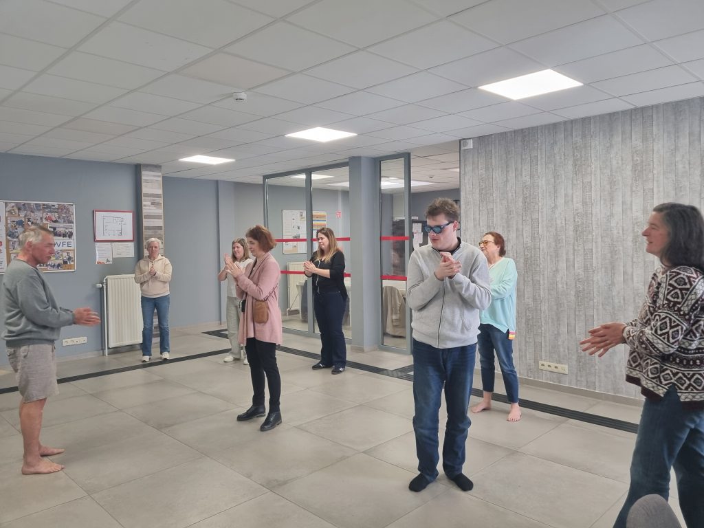 Eric est debout de profil, tourné vers le reste du groupe. Ses mains sont positionnées l'une face à l'autre devant lui.
Alexandre, face à lui, semble très concentré sur ses mains qu'il frotte l'une contre l'autre.
En arrière-plan, on voit  Rosa, Stéphanie, Katty, Delphine et Pascale participant au même geste des mains.