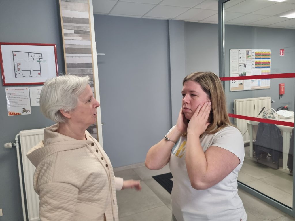 Stéphanie est en train d'exécuter un geste de massage sur son visage. Ses deux mains sont posées sur ses mâchoires, sous les oreilles, et elle applique une pression. Ses yeux sont ouverts, et elle semble écouter Rosa tout en se concentrant sur le geste et la sensation.