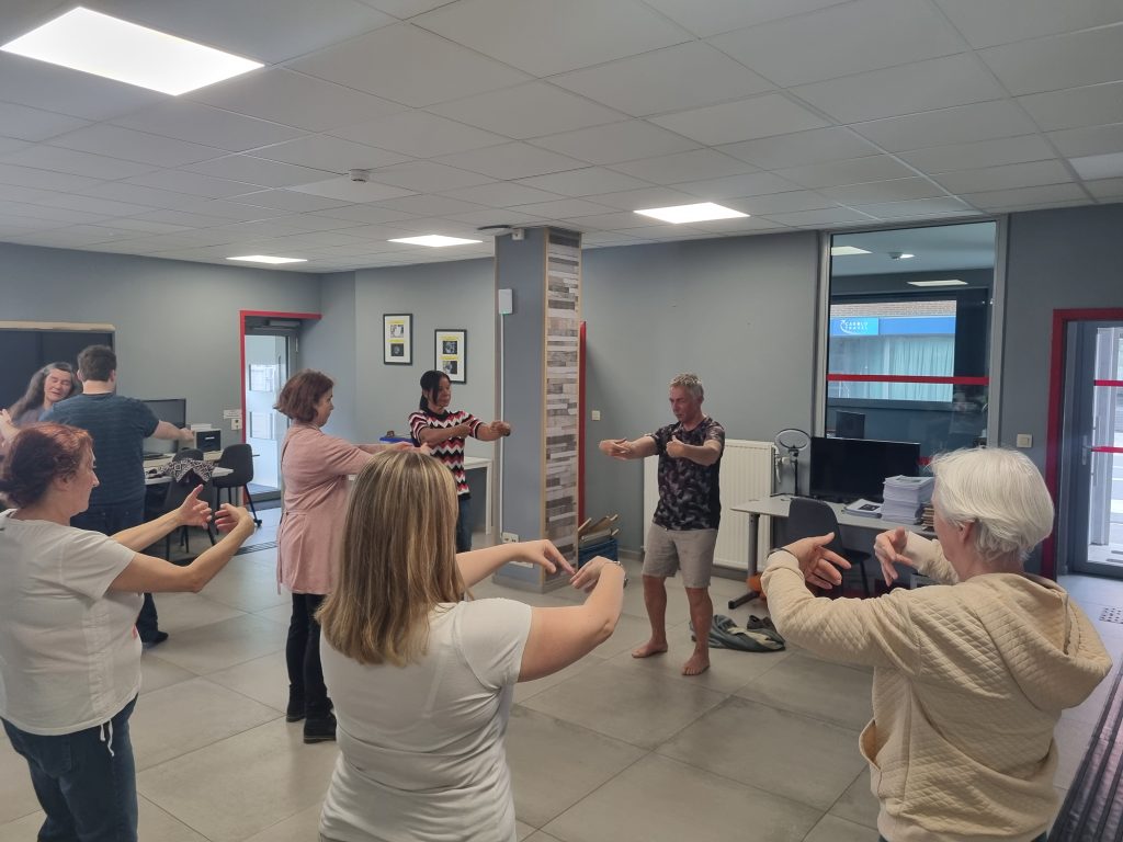 Cette image montre à nouveau le groupe réuni dans la salle polyvalente, cette fois-ci engagé dans un exercice de groupe coordonné. Tout le monde semble effectuer le même mouvement, souvent appelé « embrasser l'arbre » dans la pratique du Qi Gong.