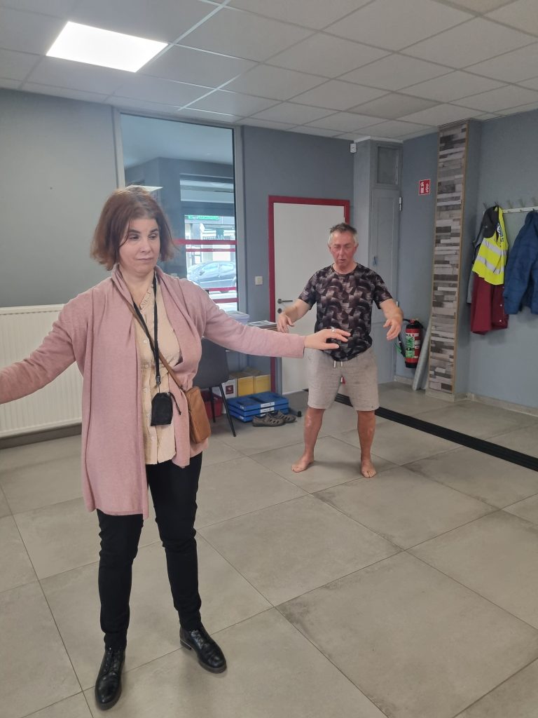 Katty et Eric occupent l'espace central. Elle est en pleine action, le corps tendu vers la gauche. Ses bras sont largement ouverts pour maintenir l'équilibre, avec ses paumes face au sol. Katty regarde attentivement vers le bas, concentrée sur sa posture, comme si elle marchait sur un fil imaginaire. Eric est positionné derrière Katty. Il adopte une posture stable, les jambes écartées et fléchies. Ses bras sont arrondis devant son buste, dans un geste de concentration.